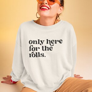 May include: White sweatshirt with the text "only here for the rolls." printed in black. The model is wearing the sweatshirt with brown pants and orange sunglasses.