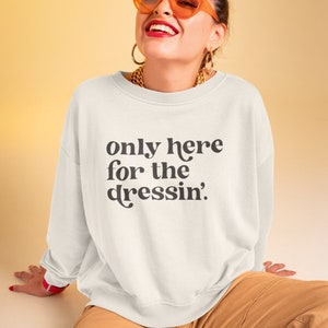 May include: White sweatshirt with the text "only here for the dressin'" printed in black. The model is wearing the sweatshirt with tan cargo pants and orange sunglasses.