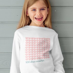 May include: A young girl wearing a white long-sleeved shirt with a pink heart pattern and the text "100 days smarter." She is wearing blue jeans and colourful trainers. She is standing in front of a grey wood panel wall.