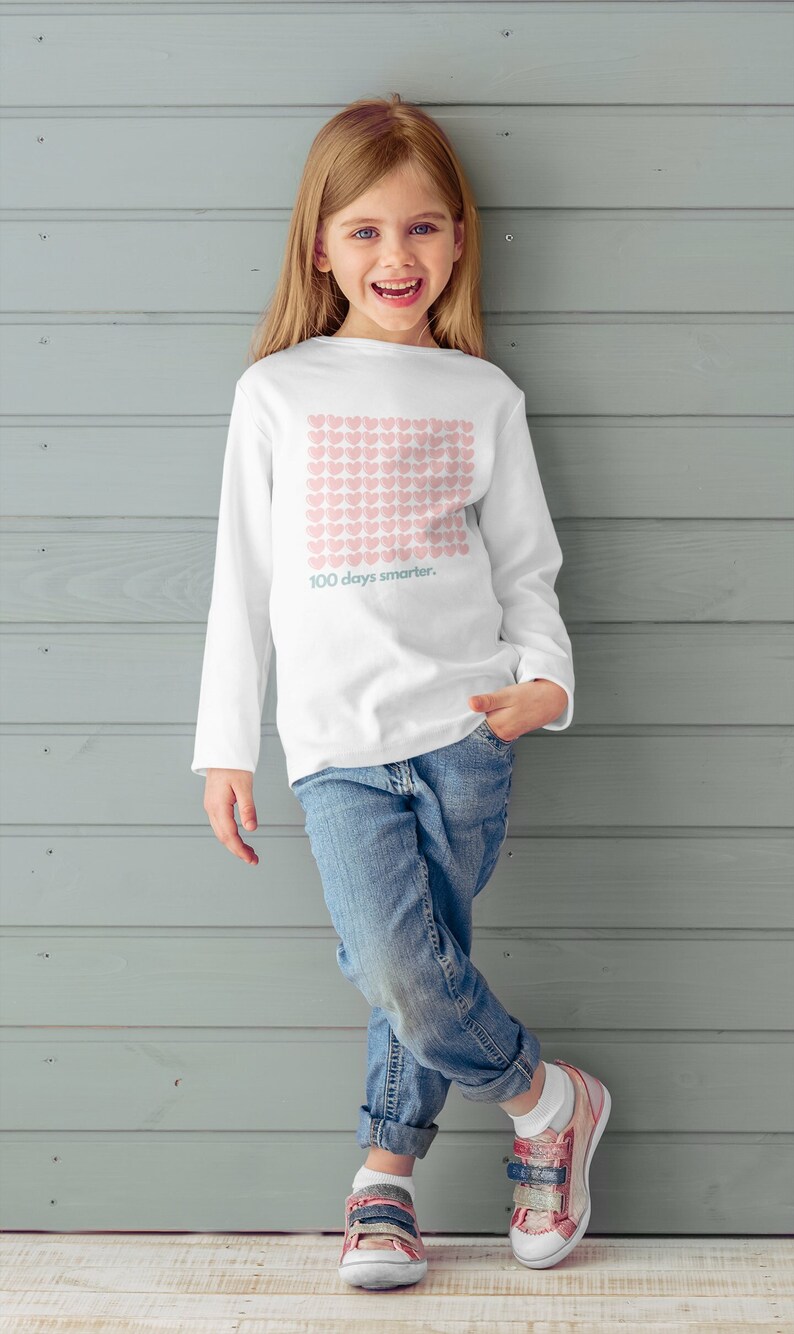 May include: A young girl wearing a white long-sleeved shirt with a pink heart pattern and the text "100 days smarter." She is wearing blue jeans and colourful trainers. She is standing in front of a grey wood panel wall.