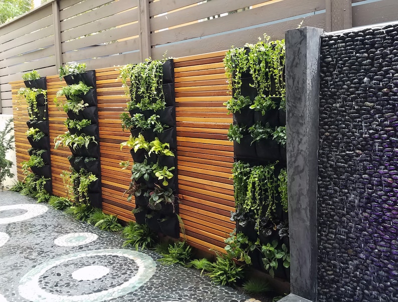 Puede incluir: Un jard&iacute;n vertical con m&uacute;ltiples bolsillos de tela negra llenos de plantas verdes, montado en una valla de madera. El jard&iacute;n est&aacute; al lado de una pared de piedra negra con una fuente.