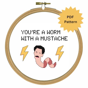 May include: A white embroidery hoop with a brown wooden frame. The hoop contains a cross-stitch pattern that says "YOU'RE A WORM WITH A MUSTACHE" with a cartoon worm with a mustache and lightning bolts on either side.