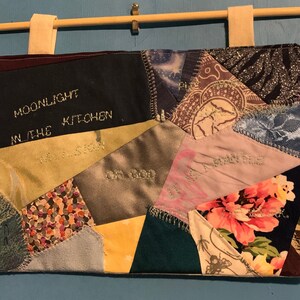 May include: A patchwork wall hanging with various fabric pieces in shades of blue, green, gold, and floral patterns. Embroidered text includes "MOONLIGHT IN THE KITCHEN" and other phrases. Beige fabric loops are attached for hanging.