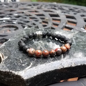 May include: A beaded bracelet featuring black lava rock beads and reddish-brown gemstone beads. The bracelet includes a silver-colored metal accent bead. The bracelet is displayed on a dark gray stone surface.