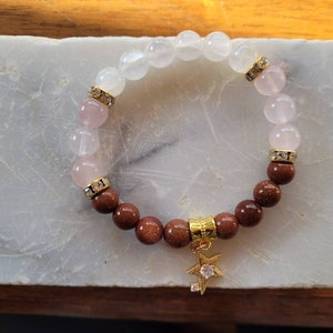 Könnte beinhalten: Ein braunes und rosafarbenes Edelstein-Armband mit einem goldenen Stern-Anhänger. Das Armband besteht aus runden Perlen und hat einen goldenen Verschluss.