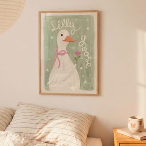 May include: A framed art print featuring a whimsical illustration of a white goose with a pink bow and a flower. The words "Silly Goose" are written in cursive on a light green background. The artwork is in a wooden frame.