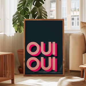 May include: A rectangular framed wall art piece featuring a dark charcoal background with the word OUI repeated twice in a stacked layout. The letters are rendered in a bold, layered pink and light coral typography with a distinct drop shadow effect.
