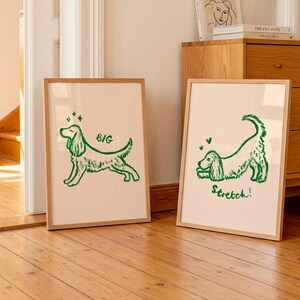 May include: Two framed art prints with a light beige background and a green dog illustration. One print says "BIG" and the other says "Stretch!". The prints are in wooden frames and are displayed in a room with wooden floors.