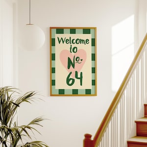 May include: A framed print with the words "Welcome to No. 64" in green and pink. The artwork has a pink heart and a cream background with a green checkered border. The frame is a warm gold colour.