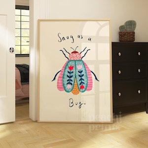 Peut inclure: Une affiche encadrée avec une illustration colorée d'un insecte stylisé et le texte "Snug as a Bug". Le motif de l'insecte comprend des éléments roses, bleus et oranges. L'affiche est encadrée dans un cadre en bois clair.