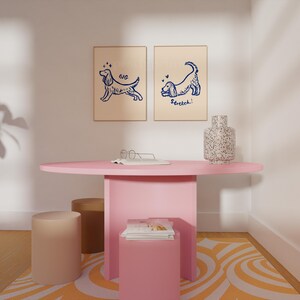 May include: A pink oval table with a matching pink base, two beige cylindrical stools, and a small pink square table. Two framed dog illustrations hang on the wall. A speckled vase sits on the table. The rug has an orange and white pattern.