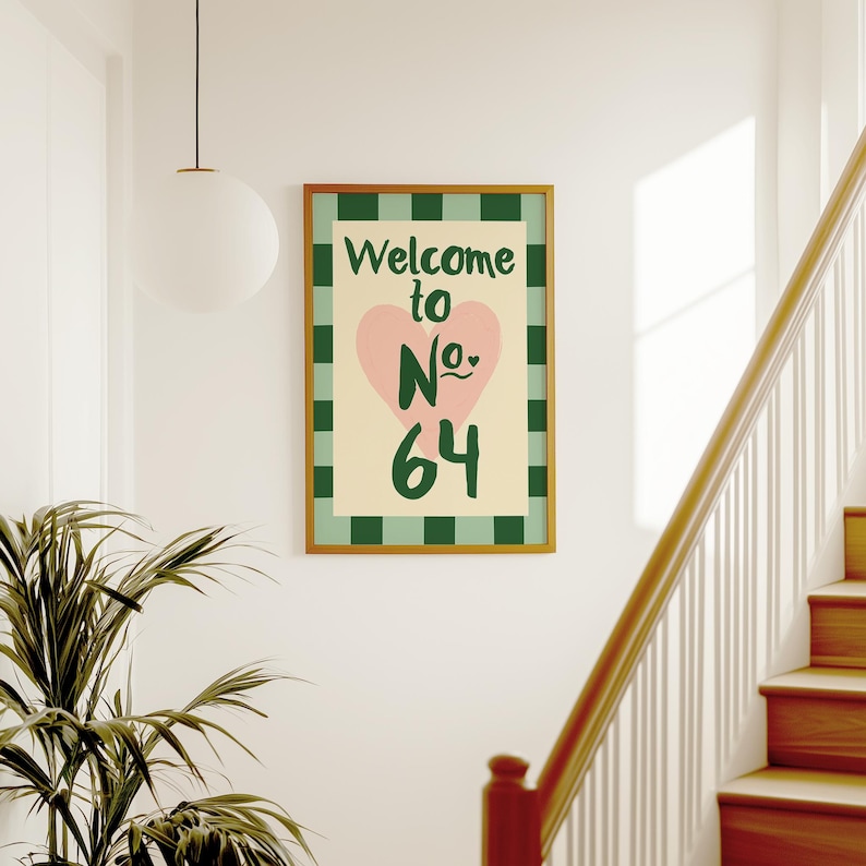May include: A framed print with the words "Welcome to No. 64" in green and pink. The artwork has a pink heart and a cream background with a green checkered border. The frame is a warm gold colour.