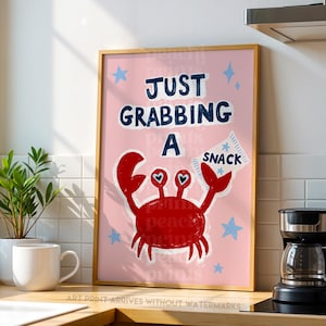 May include: A framed art print with a light pink background featuring a red crab with heart-shaped eyes holding a white snack bag. The text reads JUST GRABBING A SNACK in dark blue letters, surrounded by small light blue stars on the print.