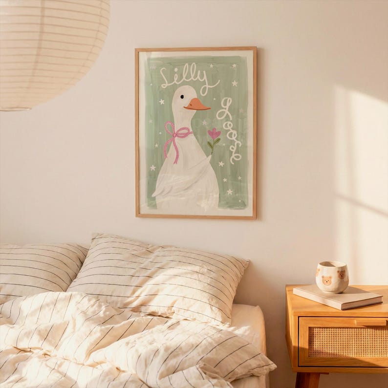 May include: A framed art print featuring a whimsical illustration of a white goose with a pink bow and a flower. The words "Silly Goose" are written in cursive on a light green background. The artwork is in a wooden frame.
