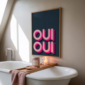 May include: A rectangular wall art print in a light wood frame featuring the text OUI OUI in bold, layered pink and light pink typography against a dark navy blue background. The artwork is displayed above a white bathtub with a wooden tray.
