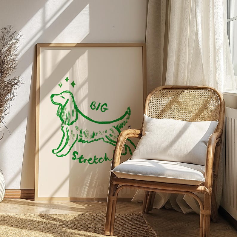 May include: A framed art print with a green dog illustration and the words "BIG Stretch". The artwork is set against a cream backdrop. A woven chair with a white cushion and pillow is in the foreground.