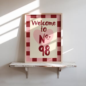 May include: A framed print with the words "Welcome to No. 98" in burgundy script. A pink heart is centered below the words. The print has a pink and burgundy checkered border and is displayed on a white shelf.