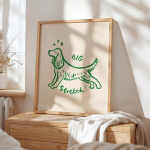 May include: A framed art print featuring a green line drawing of a dog in a stretching pose. The words "BIG Stretch!" are written in green below the dog. The background is a light beige color, and the frame is made of light wood.