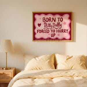 May include: A framed wall art print with the text "BORN TO Dilly Dally FORCED TO HURRY UP" in pink and red. The artwork is displayed above a bed with yellow bedding and pillows. A gold lamp sits on a wooden nightstand.