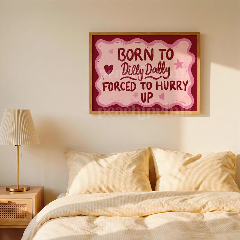 May include: A framed wall art print with the text "BORN TO Dilly Dally FORCED TO HURRY UP" in pink and red. The artwork is displayed above a bed with yellow bedding and pillows. A gold lamp sits on a wooden nightstand.