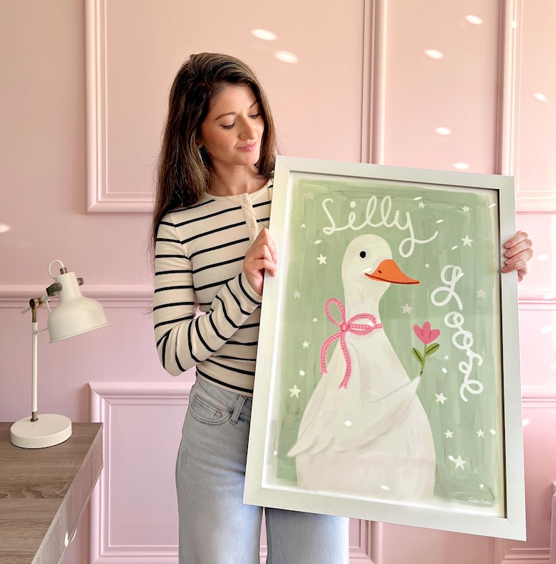 May include: A framed art print featuring a white goose with an orange beak on a sage green background. The goose wears a pink bow and holds a pink flower. The text "Silly Goose" is written in white script, surrounded by small white stars.
