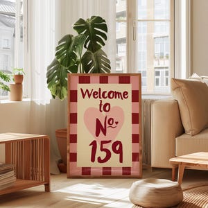 May include: A framed sign with the text "Welcome to No. 159". The design includes a pink heart and a red and pink checked border. The sign is in a wooden frame, creating a welcoming feel.