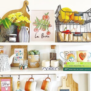 May include: A white shelf unit with various items, including a peach-themed print with the text "La Peche", a framed print with the text "yes girl you can", a succulent in a ceramic pot with a cheetah print, two mugs with colorful designs, and two wire baskets labeled "FRUIT" and "VEGETABLES".
