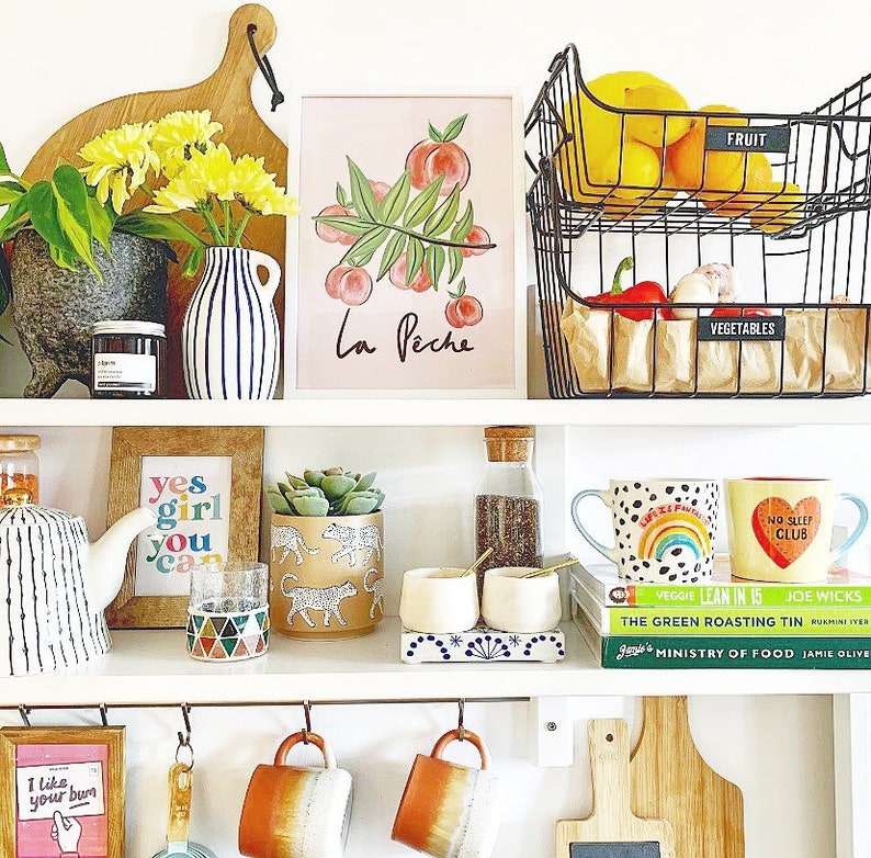 May include: A white shelf unit with various items, including a peach-themed print with the text "La Peche", a framed print with the text "yes girl you can", a succulent in a ceramic pot with a cheetah print, two mugs with colorful designs, and two wire baskets labeled "FRUIT" and "VEGETABLES".