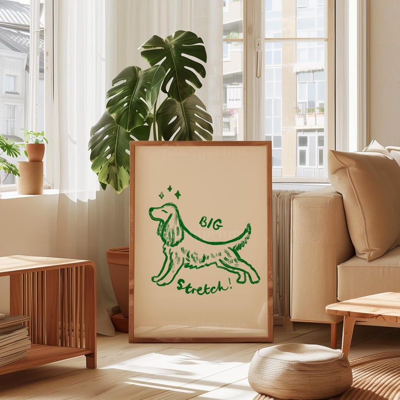 May include: A framed print featuring a green illustration of a dog with the words "BIG Stretch!" on a cream background. The artwork is in a wooden frame and is displayed in a well-lit room with a large plant and a sofa.