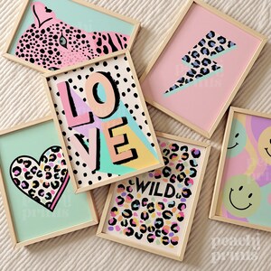 May include: A collection of framed art prints with a playful, modern aesthetic. Designs include a pink leopard, a lightning bolt, a heart, the word "LOVE", the word "WILD", and smiley faces. Each print has a wooden frame and a colorful background.