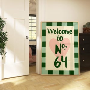 May include: A framed print with a green and white checked border. The artwork displays the text "Welcome to No. 64" in green, with a pink heart. The print is in a room with a black chest of drawers and a potted plant.