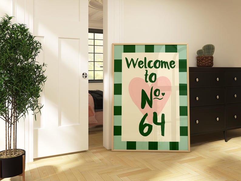 May include: A framed print with a green and white checked border. The artwork displays the text "Welcome to No. 64" in green, with a pink heart. The print is in a room with a black chest of drawers and a potted plant.