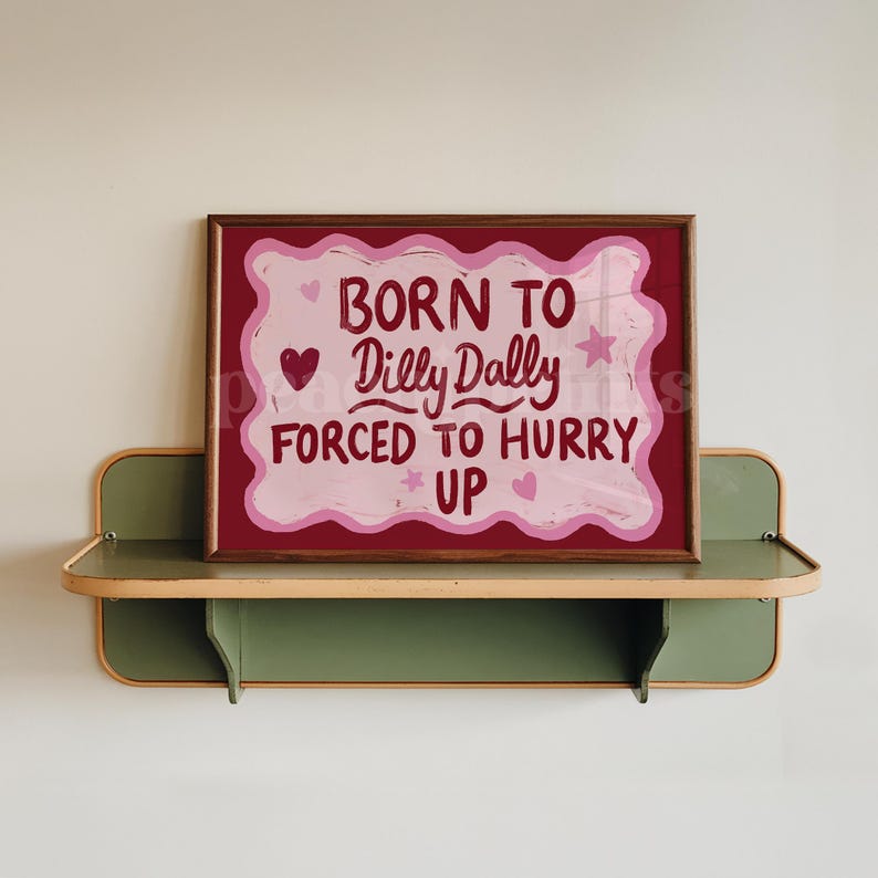 May include: A framed art print with the text "BORN TO DILLY DALLY FORCED TO HURRY UP" in a playful font. The artwork features a maroon background with a pink cloud-like shape and decorative hearts and stars. The frame is brown.