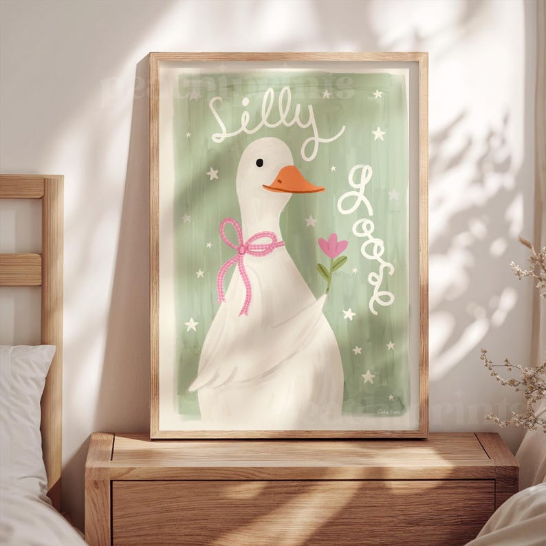 May include: A framed wall art print featuring a white goose with an orange beak, wearing a pink ribbon bow around its neck and holding a small pink flower. The background is sage green with white stars and the text Silly Goose in white script.