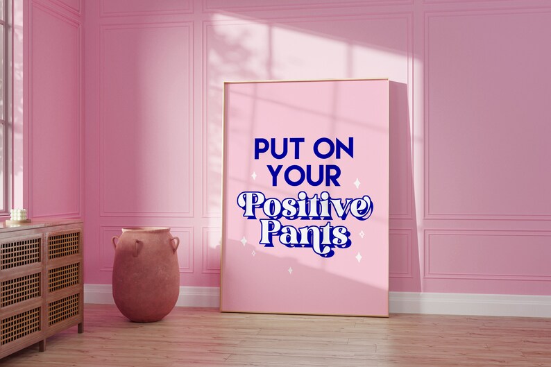 May include: A pink framed poster with the text "PUT ON YOUR Positive Pants" in blue and white lettering with white stars on a pink background.