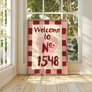 May include: Framed wall art with the words "Welcome to No. 1548" in burgundy script. A pink heart is centered on the cream-coloured background. The frame has a pink and burgundy chequered pattern.