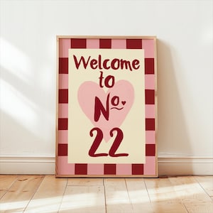 May include: A framed print with the text "Welcome to No. 22" in burgundy. The design includes a pink heart on a cream background, framed by a pink and burgundy checkered border. The frame is a light brown colour. The print is displayed on a wooden floor.