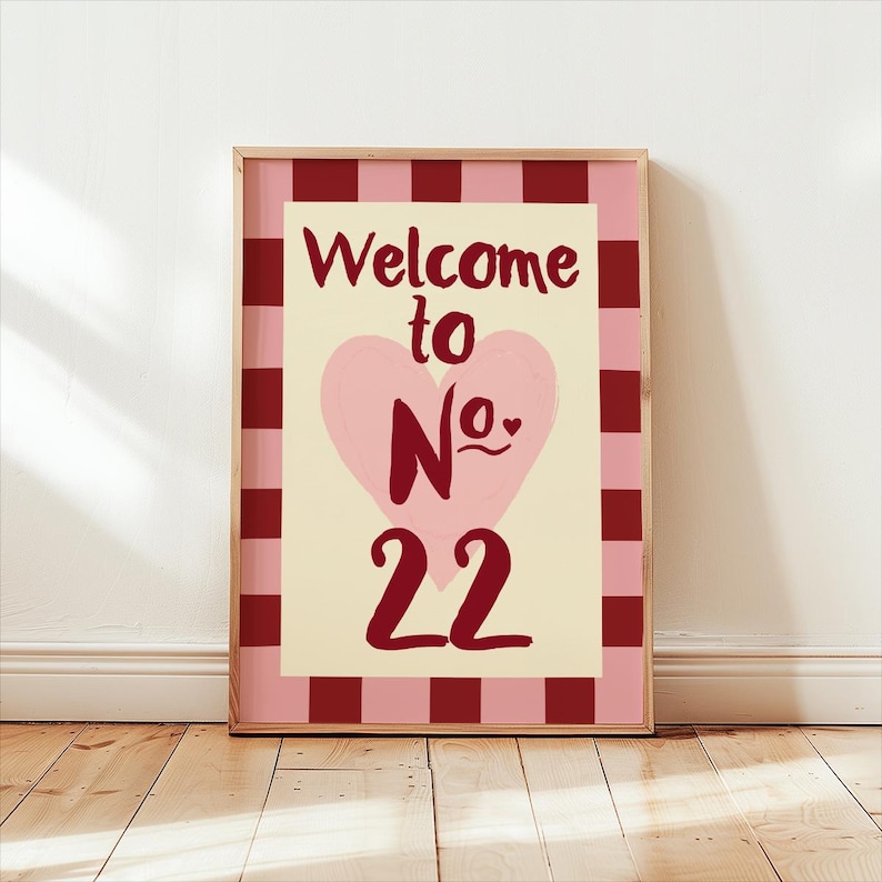 May include: A framed print with the text "Welcome to No. 22" in burgundy. The design includes a pink heart on a cream background, framed by a pink and burgundy checkered border. The frame is a light brown colour. The print is displayed on a wooden floor.