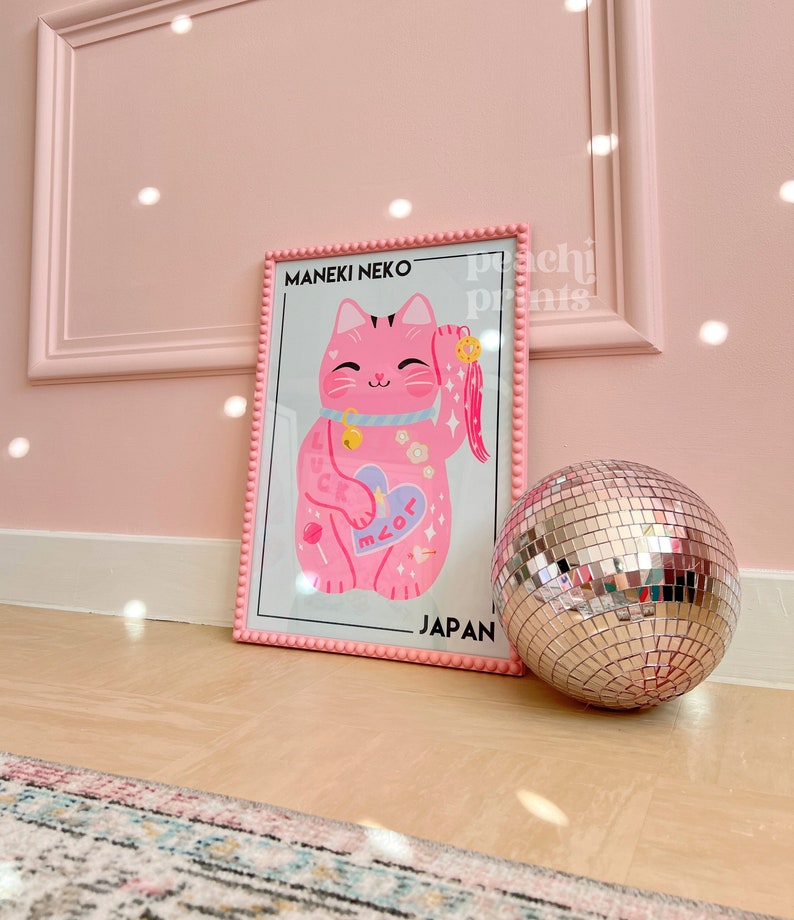 Puede incluir: Impresi&oacute;n enmarcada rosa de un Maneki Neko rosa, un gato de la suerte japon&eacute;s, con una campana y un coraz&oacute;n que dice "miau" en rosa y blanco. La impresi&oacute;n dice "Maneki Neko" y "Jap&oacute;n" en texto negro. Una bola de discoteca plateada est&aacute; en primer plano.