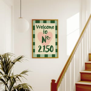 May include: A framed print with the text "Welcome to No. 2150". The design includes a pink heart and green and white checkered pattern. The artwork is set in a brown frame. The print is displayed on a white wall, near a staircase.