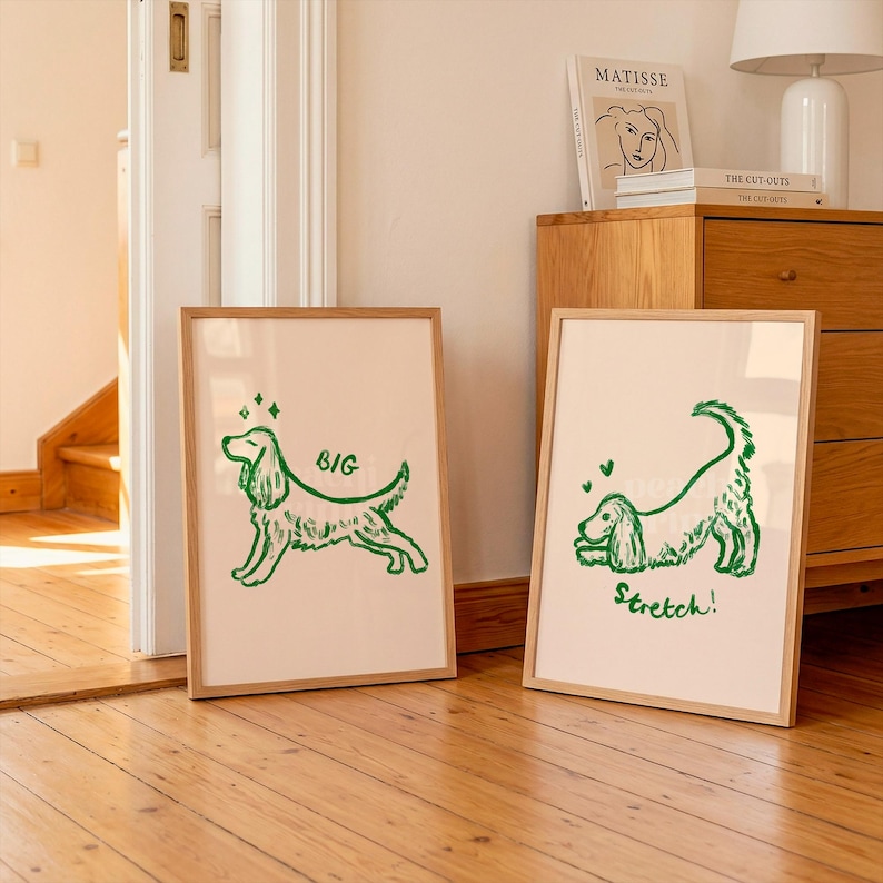 May include: Two framed art prints with a light beige background and a green dog illustration. One print says "BIG" and the other says "Stretch!". The prints are in wooden frames and are displayed in a room with wooden floors.