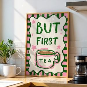 May include: A framed art print with the text "BUT FIRST TEA" in green, pink, and white. The print features a teacup illustration and star accents. The artwork is displayed on a kitchen counter with a coffee maker and a white mug.