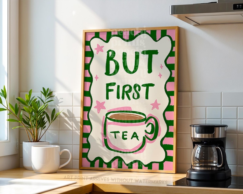 May include: A framed art print with the text "BUT FIRST TEA" in green, pink, and white. The print features a teacup illustration and star accents. The artwork is displayed on a kitchen counter with a coffee maker and a white mug.