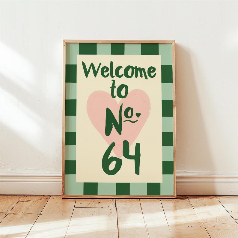 May include: A framed art print featuring the text "Welcome to No. 64" in green. A pink heart is in the center. The background is cream-colored, with a green and white checkered border. The print is in a wooden frame.