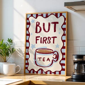 May include: A framed art print with the words "BUT FIRST TEA" in a whimsical design. The print features a red and white border, blue stars, and a coffee cup illustration. The artwork is displayed on a kitchen counter with a coffee maker and a white mug.