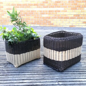 May include: Two black and white woven baskets with a striped pattern. The smaller basket is filled with green plants. The baskets are on a wooden surface.