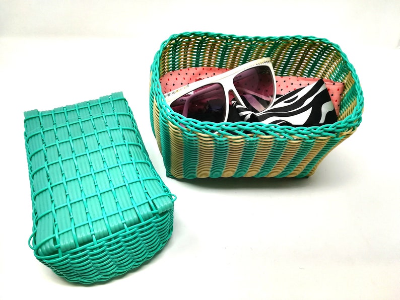 May include: Two woven baskets, one teal and one with tan and teal stripes. The striped basket is filled with a pink and white patterned scarf, a black and white zebra patterned scarf, and a pair of white sunglasses with pink lenses.