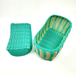 May include: Two woven baskets, one rectangular and one curved, are made of green and beige plastic. The baskets are empty and sitting on a white surface.