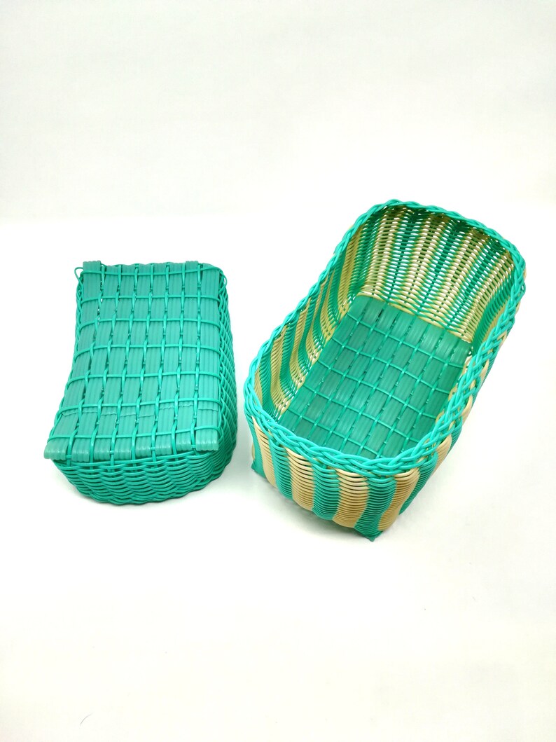 May include: Two woven baskets, one rectangular and one curved, are made of green and beige plastic. The baskets are empty and sitting on a white surface.