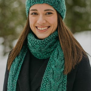 Könnte beinhalten: Ein teal-grünes Strickmützen- und Schal-Set. Die Mütze ist eine eng anliegende Beanie-Mütze, und der Schal hat ein Zopfmuster. Das Set ist aus einem weichen, strukturierten Garn gefertigt, perfekt für kaltes Wetter. Die Frau trägt einen schwarzen Mantel.